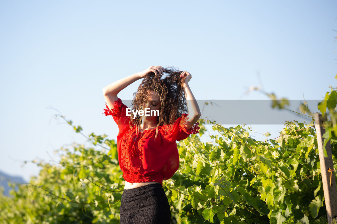 YOUNG WOMAN STANDING BY PLANT AGAINST CLEAR SKY