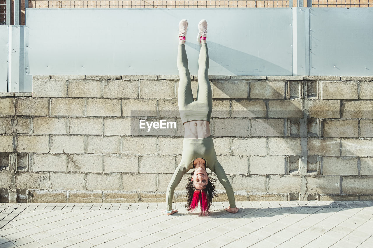 Smiling woman with hair highlights doing handstand against brick wall