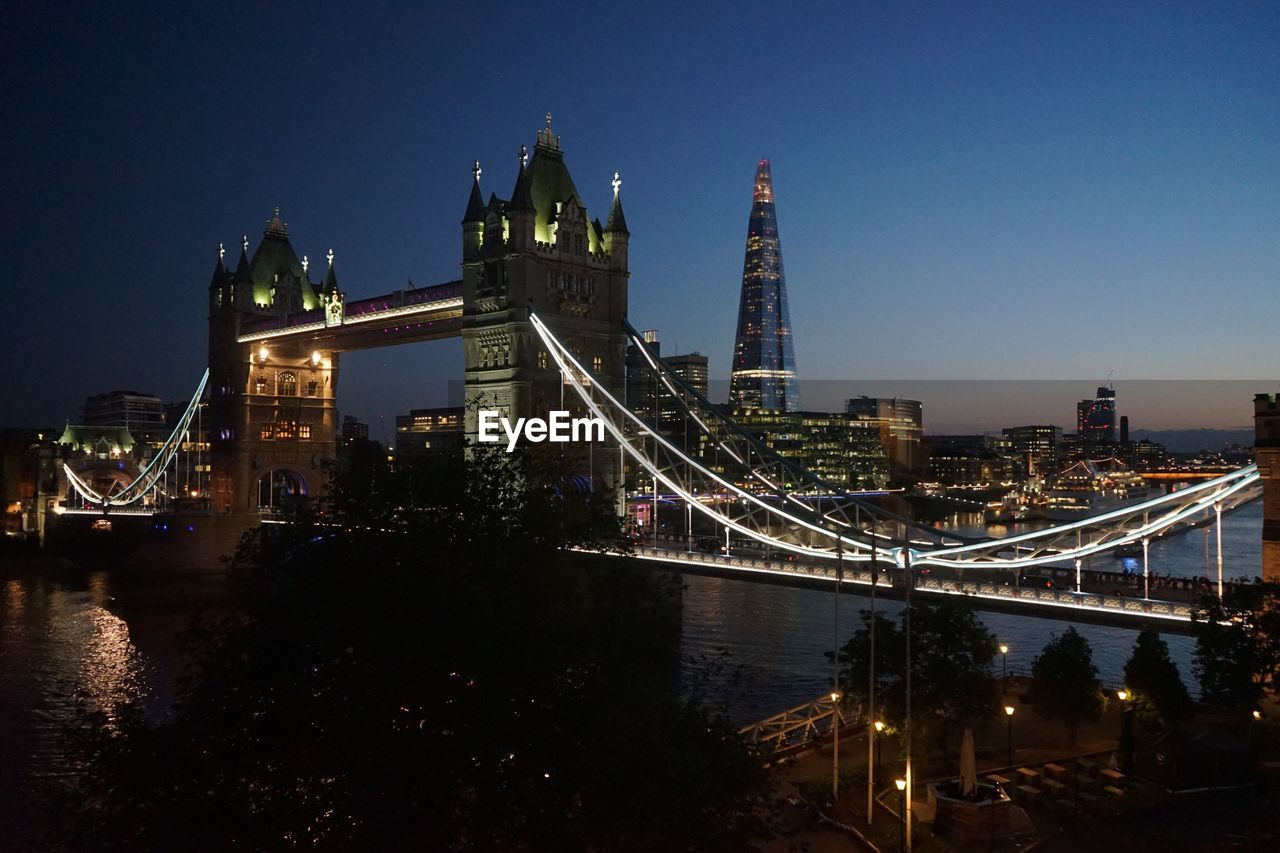 ILLUMINATED BRIDGE OVER RIVER BY BUILDINGS AGAINST SKY