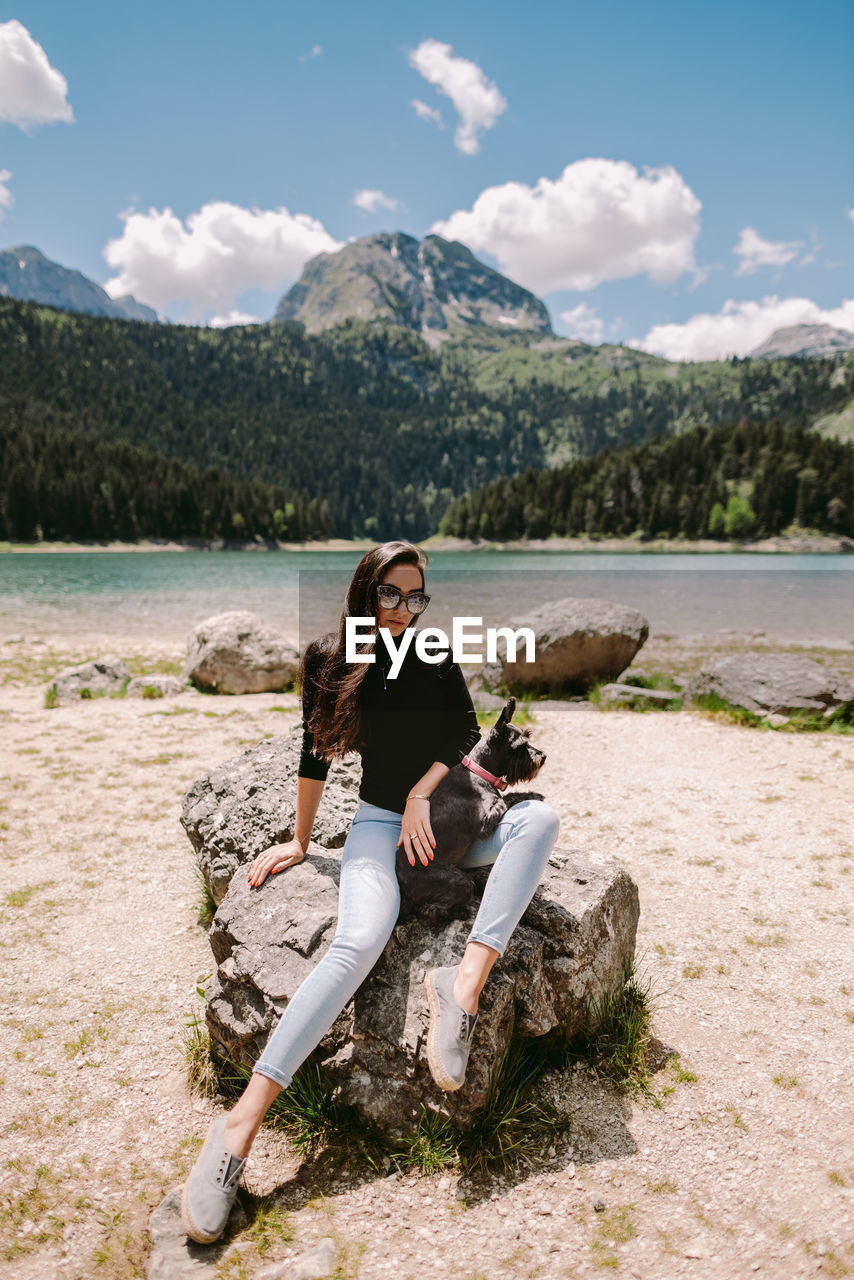 PORTRAIT OF YOUNG WOMAN SITTING ON SHORE AGAINST MOUNTAINS