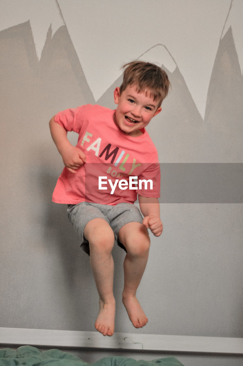 childhood, full length, child, one person, men, toddler, emotion, indoors, smiling, sitting, fun, happiness, human leg, front view, portrait, clothing, barefoot, casual clothing, person, arm, baby, hand, limb, photo shoot, cheerful, human face, lifestyles, wall - building feature, pink, looking at camera