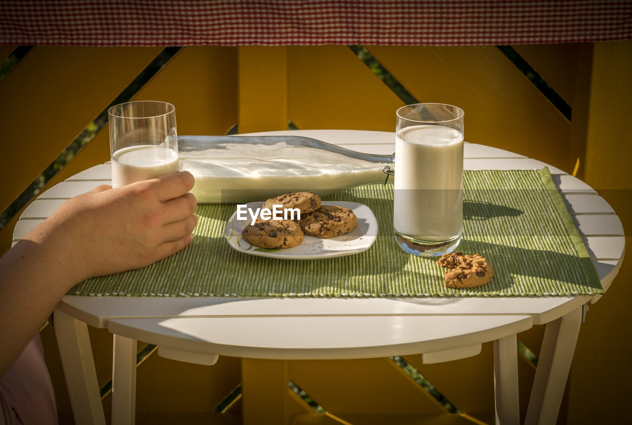 Cropped image of hand holding milk with biscuits in plate on table