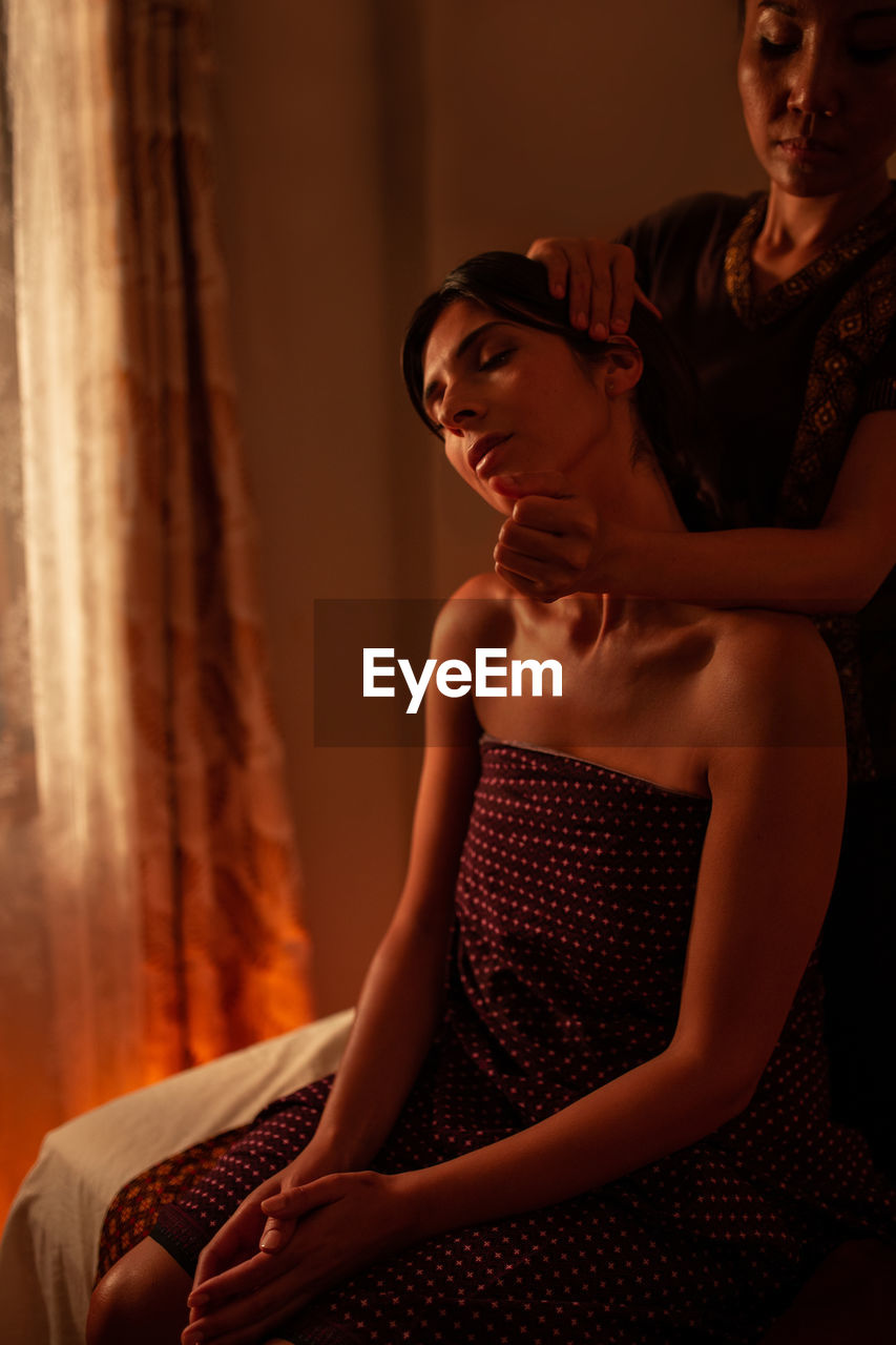 Asian female massagist in traditional clothes giving a thai massage to a caucasian woman sitting on a bed in a room lit with warm lights