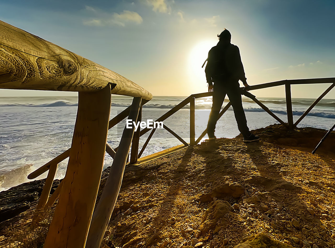 MAN STANDING ON RAILING AT BEACH AGAINST SKY