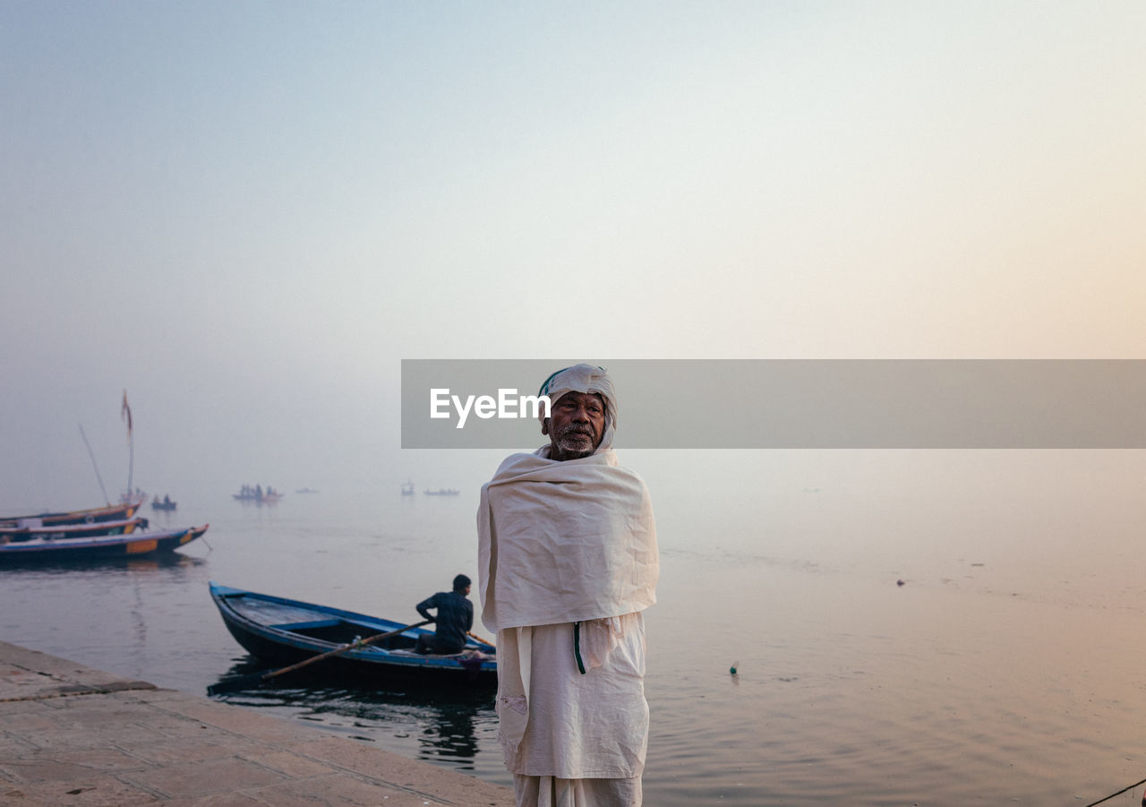 Varanasi, india - february, 2018: elderly indian man wearing traditional white clothes standing on river bank with fishing boats on water in early foggy morning