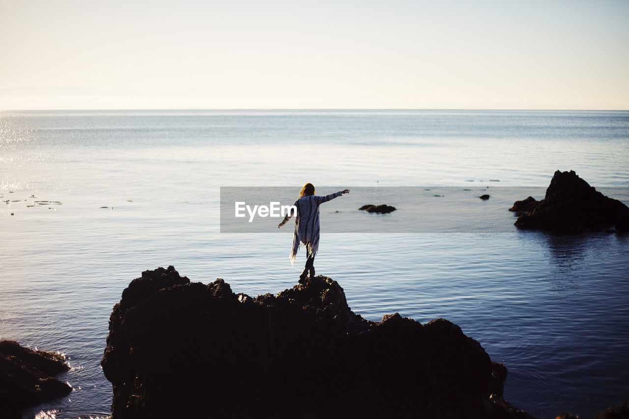 REAR VIEW OF MAN STANDING ON ROCK IN SEA