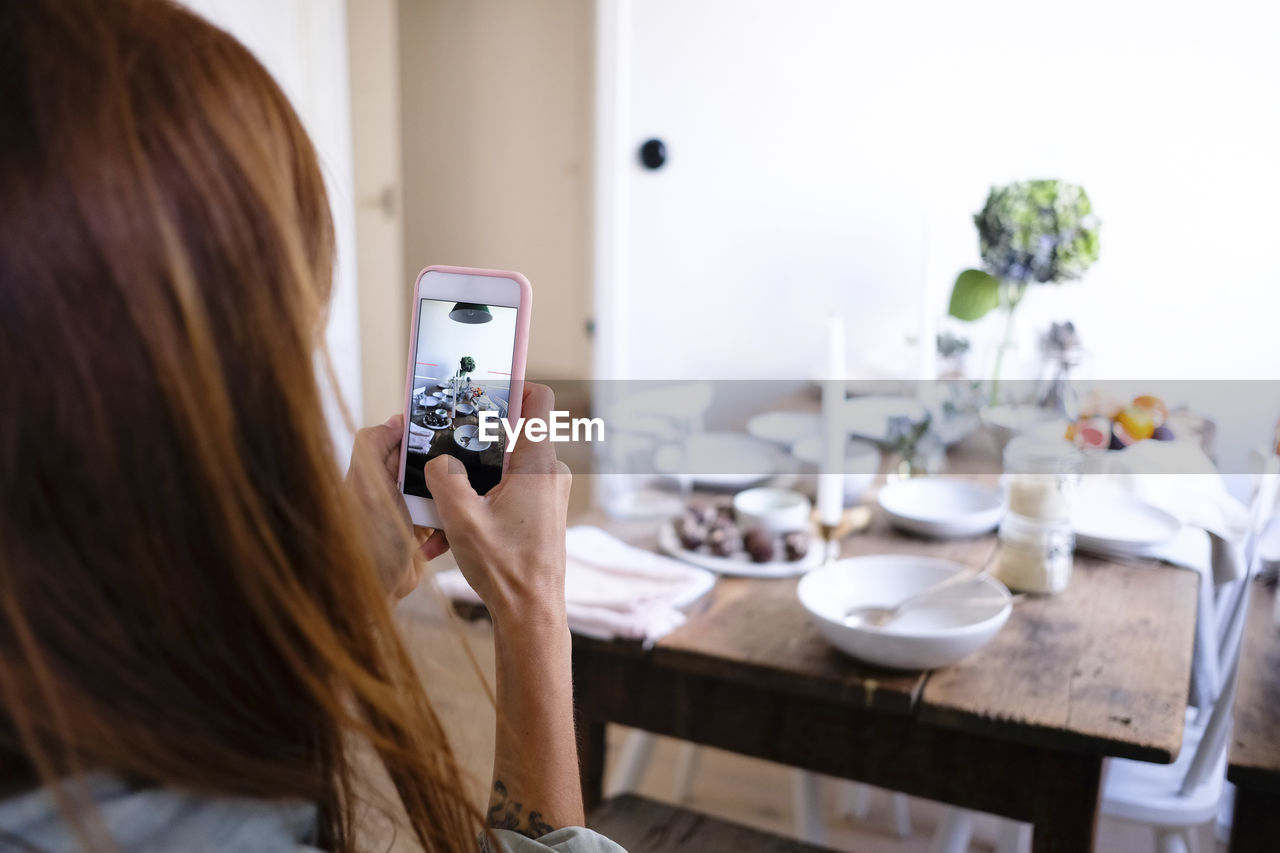 Rear view of woman photographing plates and food on table by wall at home