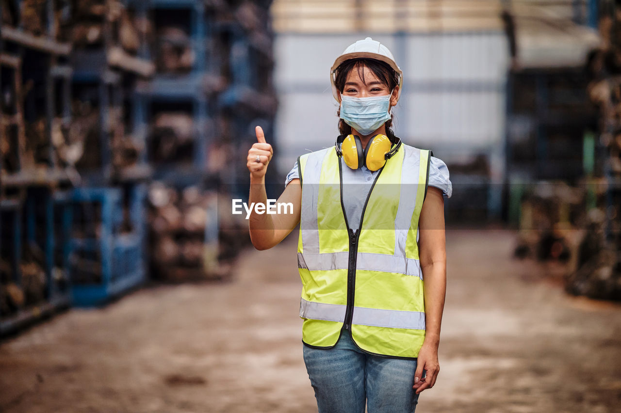 Confident female engineer wearing a mask while standing with thumbs up in a factory