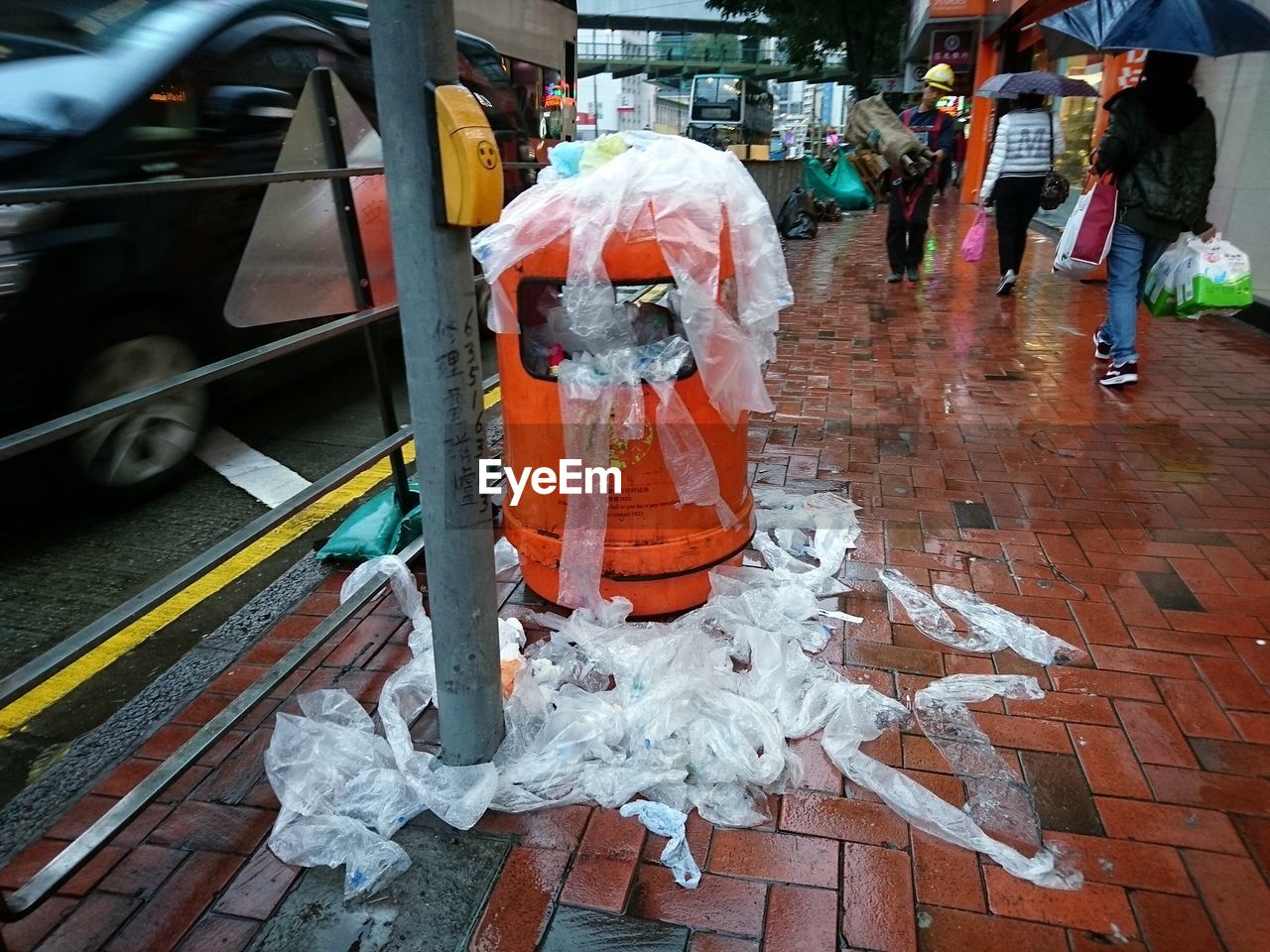 People walking by garbage bin with plastic bags on street during monsoon