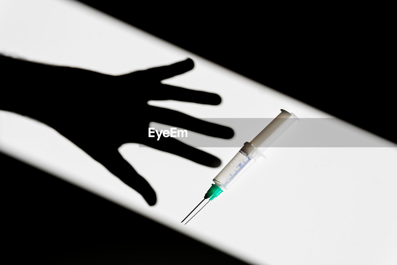 Shadow of crop anonymous person reaching out for syringe placed on white table