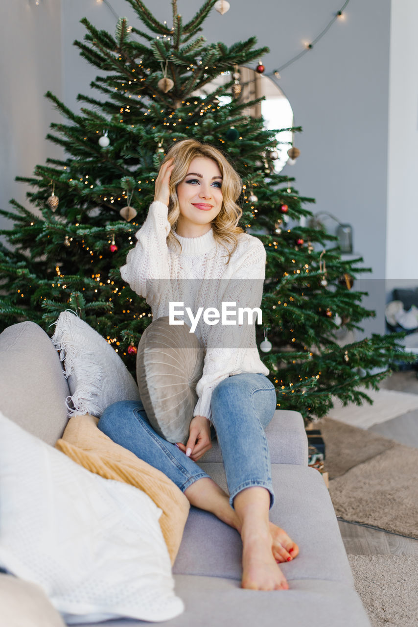 Lovey woman is sitting on a gray sofa near the christmas tree in the living room and holding 