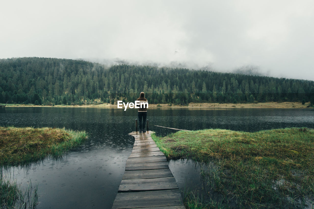 Rear view of woman standing on jetty in lake against sky during rainfall