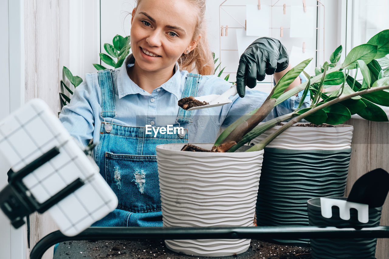 Gardener woman blogger using phone while transplants indoor plants and use a shovel on table. 