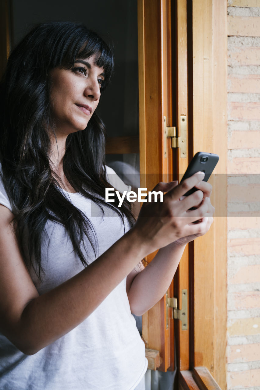 Young woman using mobile phone while standing by window at home