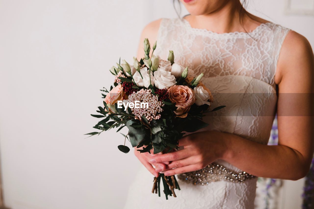Midsection of bride holding flower bouquet against wall