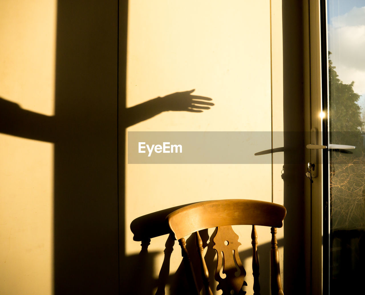 Shadow of a woman's arm and hand above a chair, against a wall next to a glass door in afternoon sun