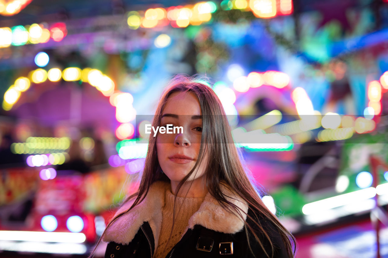 PORTRAIT OF SMILING YOUNG WOMAN STANDING AGAINST ILLUMINATED LIGHTS