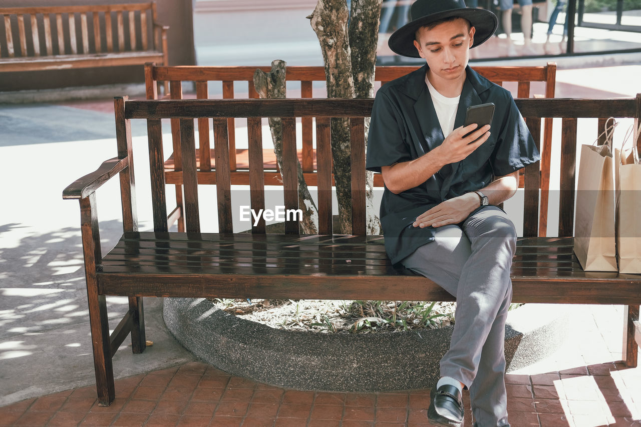 Man using mobile phone while sitting with shopping bags on bench outside store