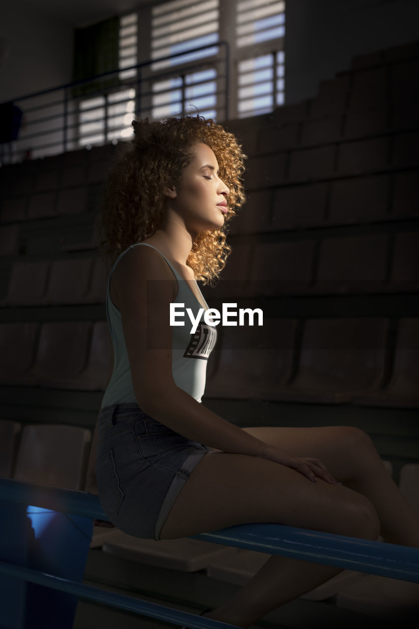 Portrait of young curly-haired woman sitting in a stadium with sunset light