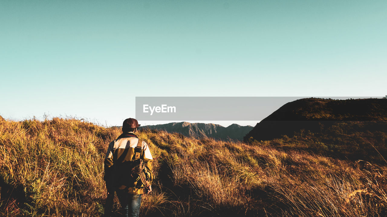 Rear view of man hiking on grassy mountain against clear sky during sunset