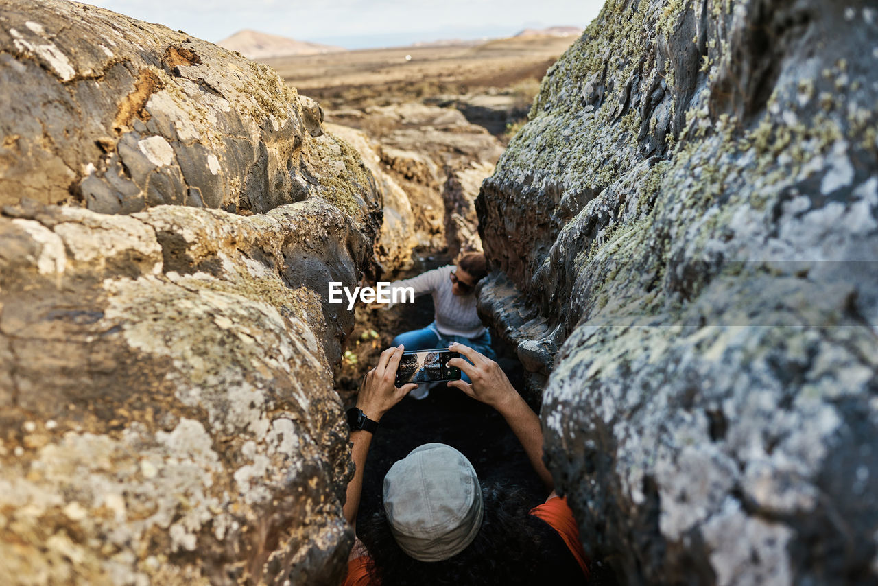 Anonymous traveler shooting friend via smartphone while standing in narrow crack between rough rocks in countryside of fuerteventura, spain