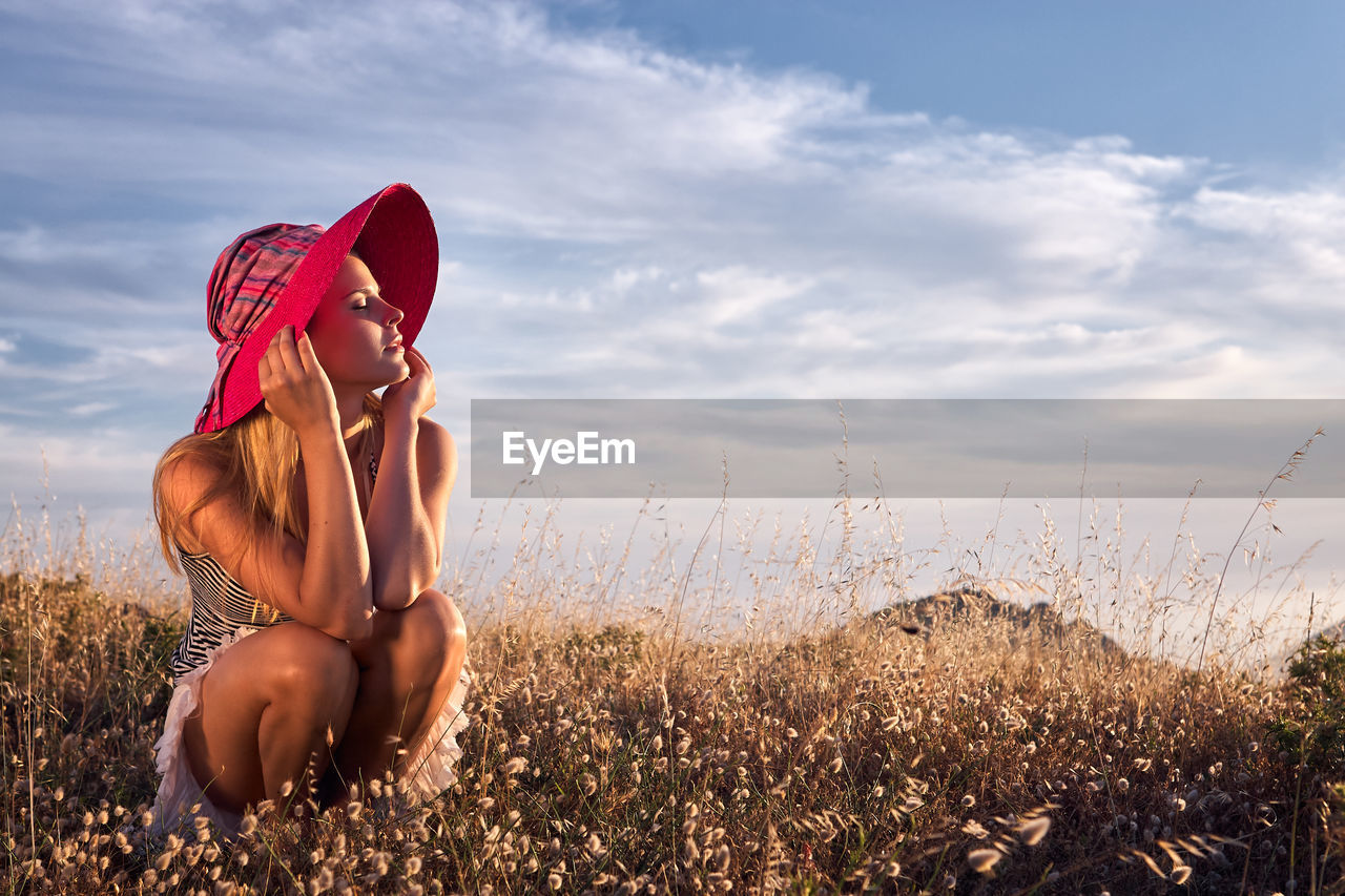 Young woman wearing hat crouching on grassy field against sky