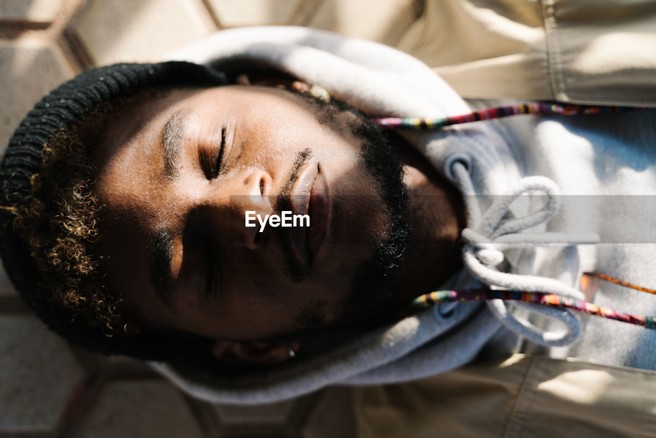 Top view of calm young bearded african american male in trendy outfit lying on ground with closed eyes on sunny day