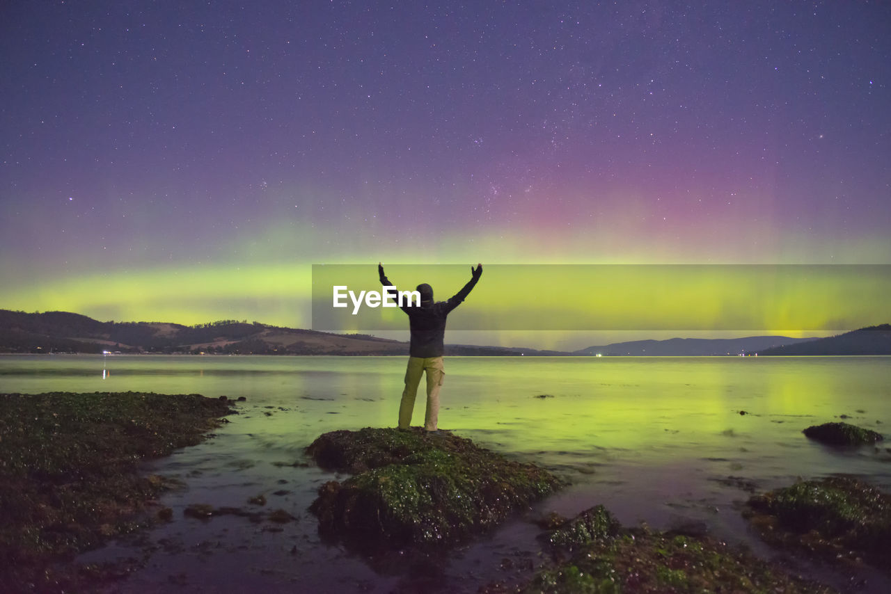 Rear view of man with arms raised standing on rock at lakeshore against northern lights at night