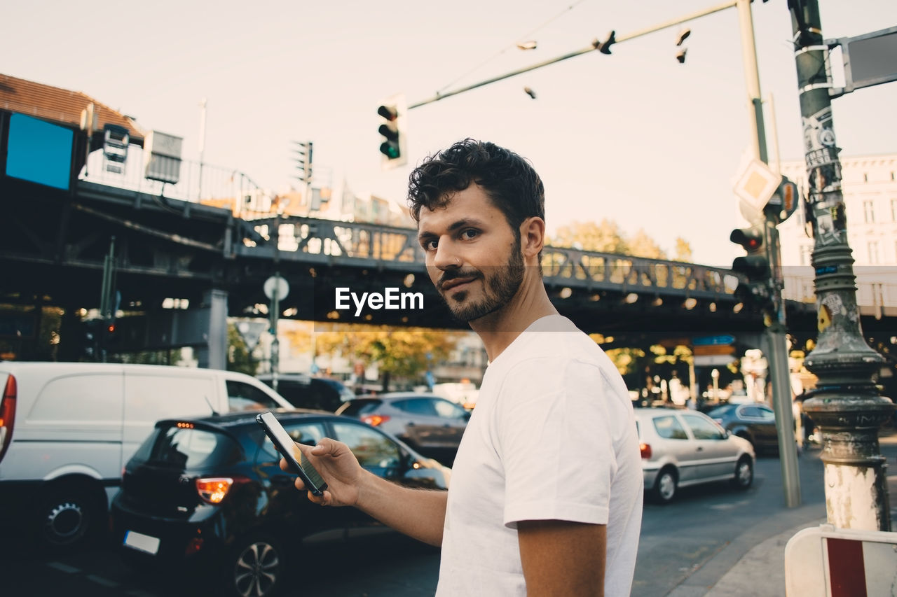 Portrait of young man holding mobile phone while standing on sidewalk in city