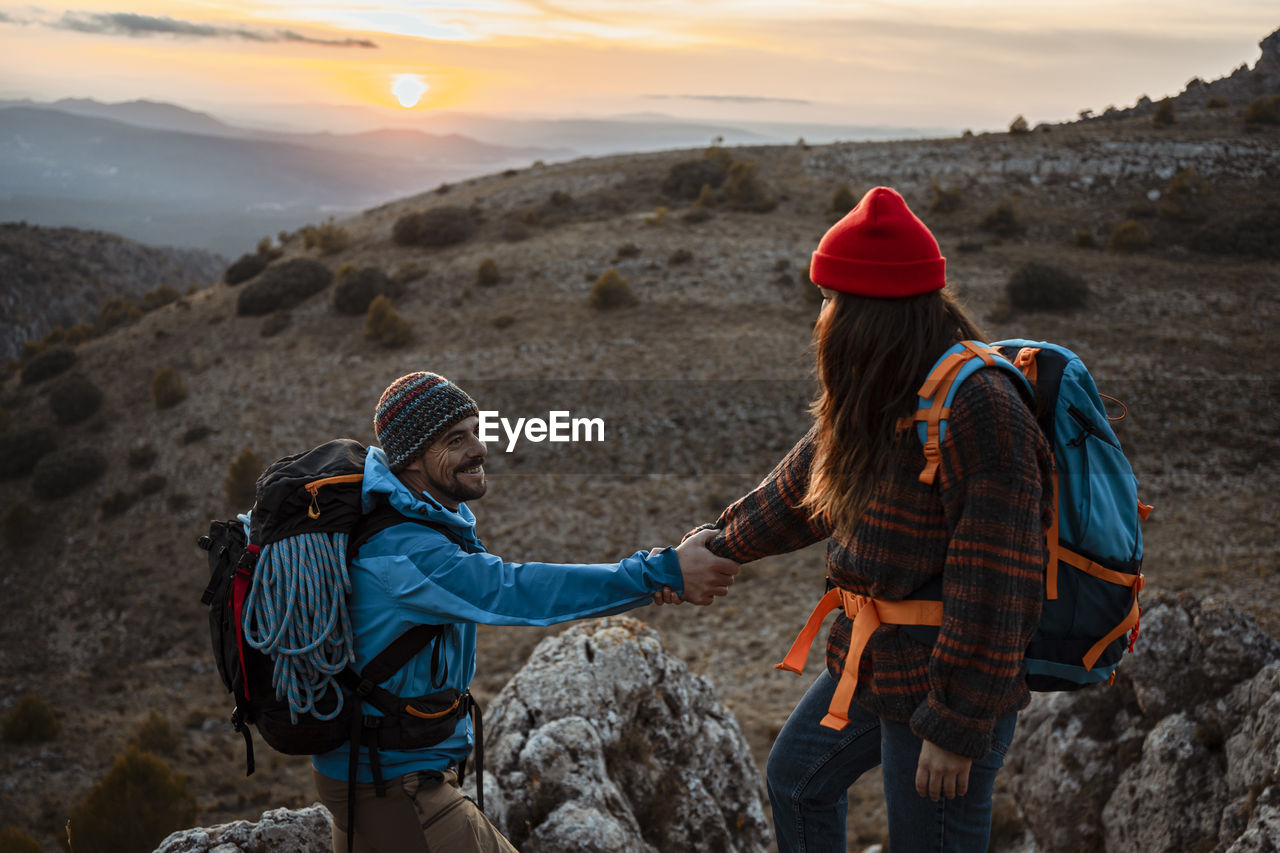 Girlfriend helping boyfriend to climb rocky mountain during sunset