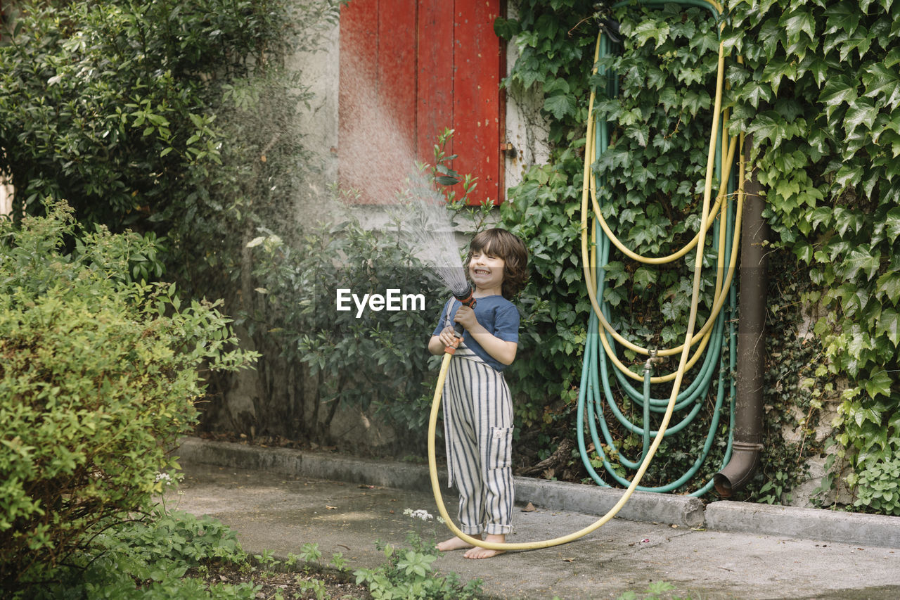 Boy enjoying holding garden hose while watering plants at backyard