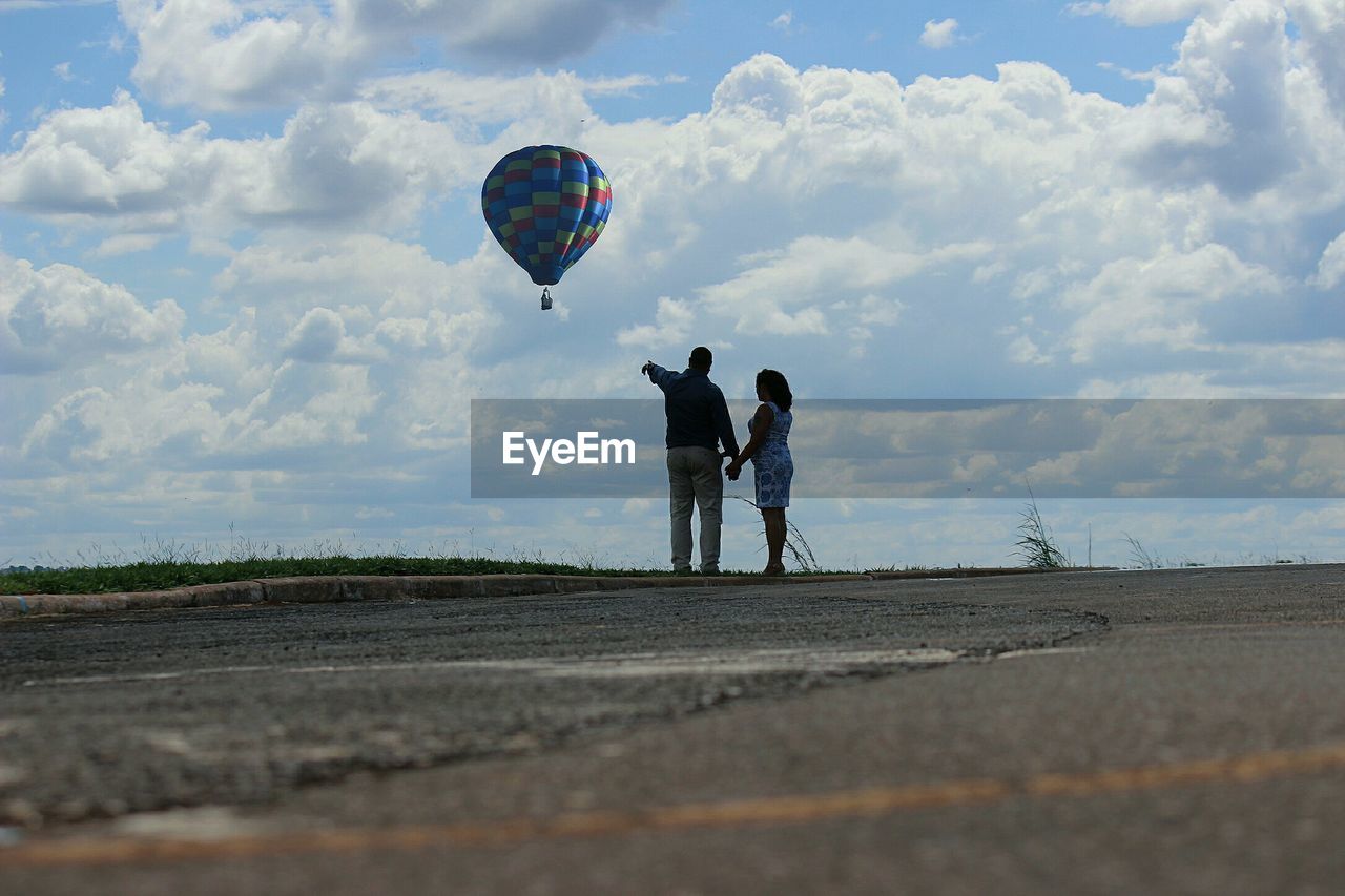 Rear view of couple standing against hot air balloon flying in cloudy sky