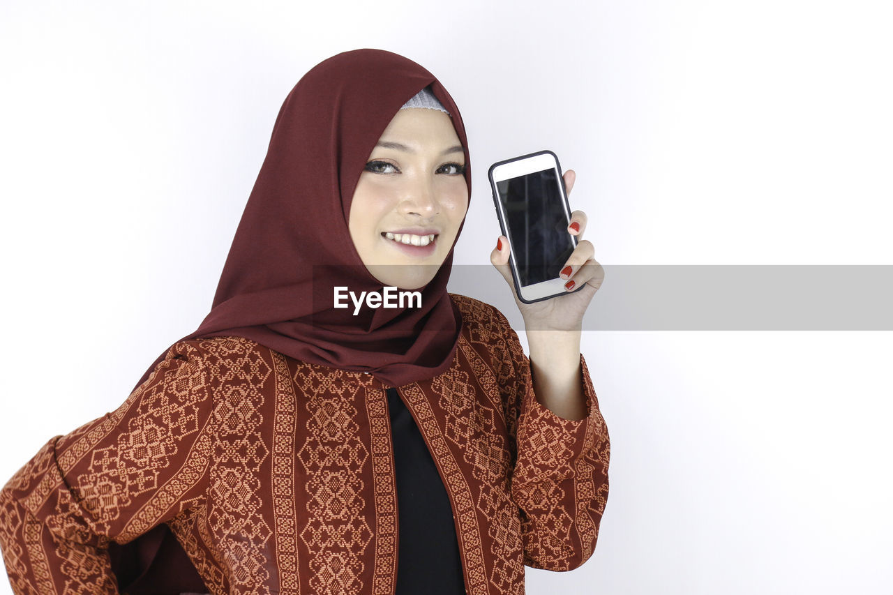 PORTRAIT OF SMILING YOUNG WOMAN USING PHONE AGAINST WHITE BACKGROUND