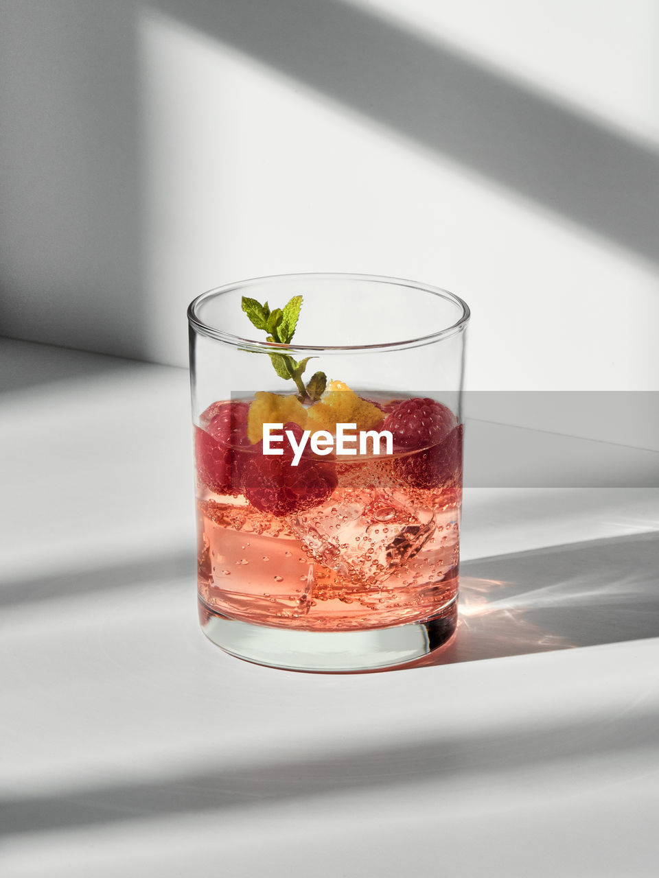 Glass with cocktail with ice cubes and raspberries with mint sprig placed on white surface
