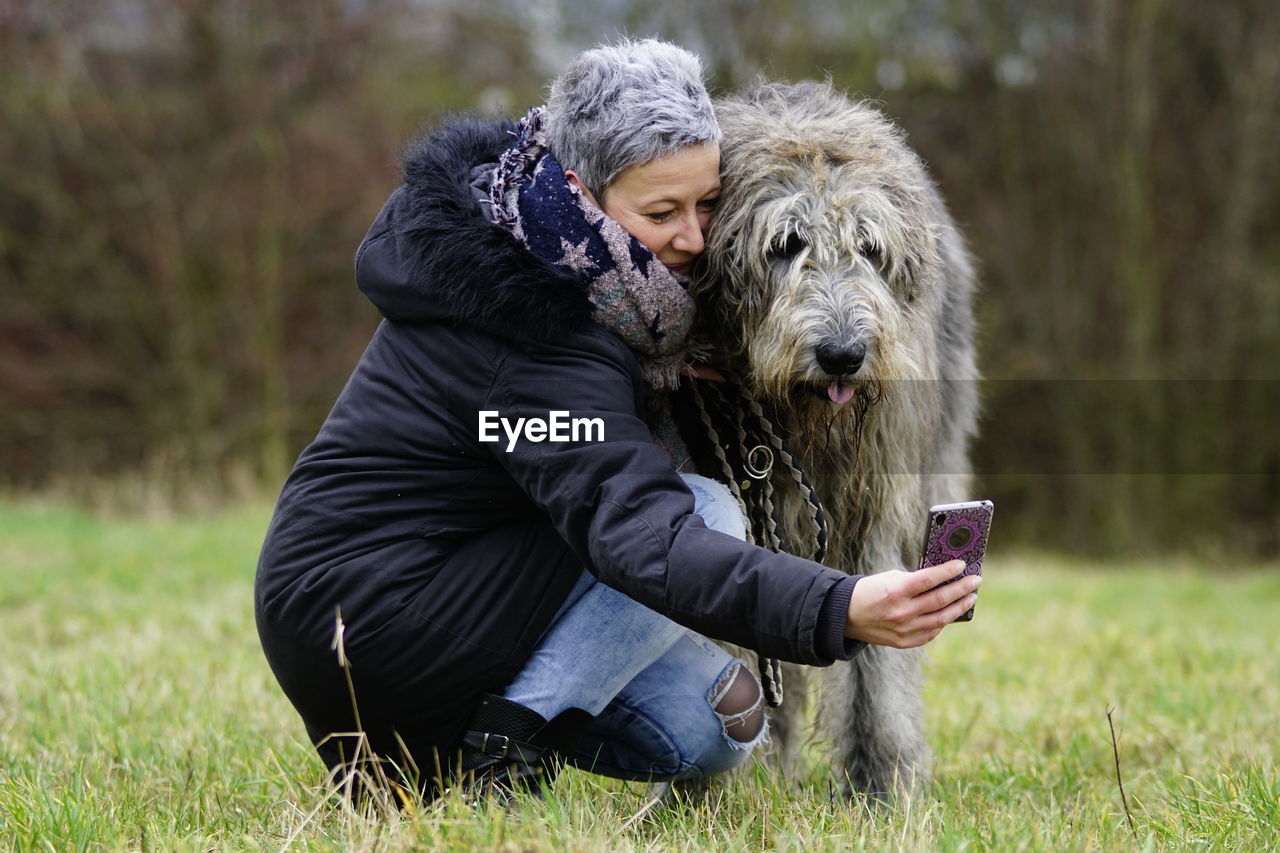 Mature woman with dog taking seflie on grassy field