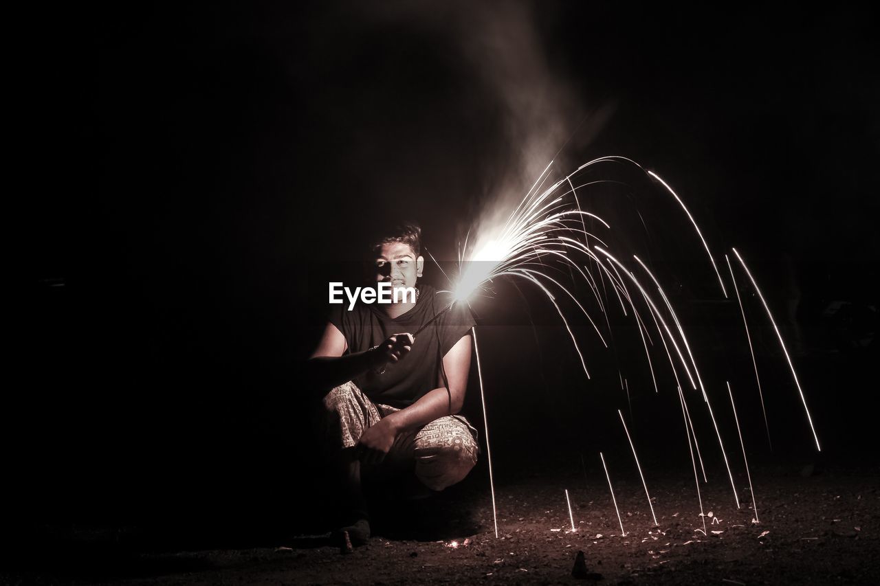 Portrait of mature man holding firework while crouching on field at night