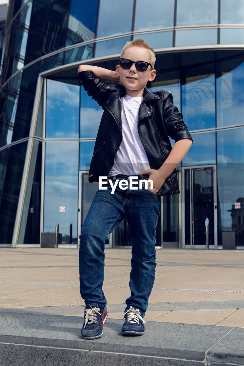 Baby boy  in sunglasses and black leather jacket smiling of the glass building in the summer,