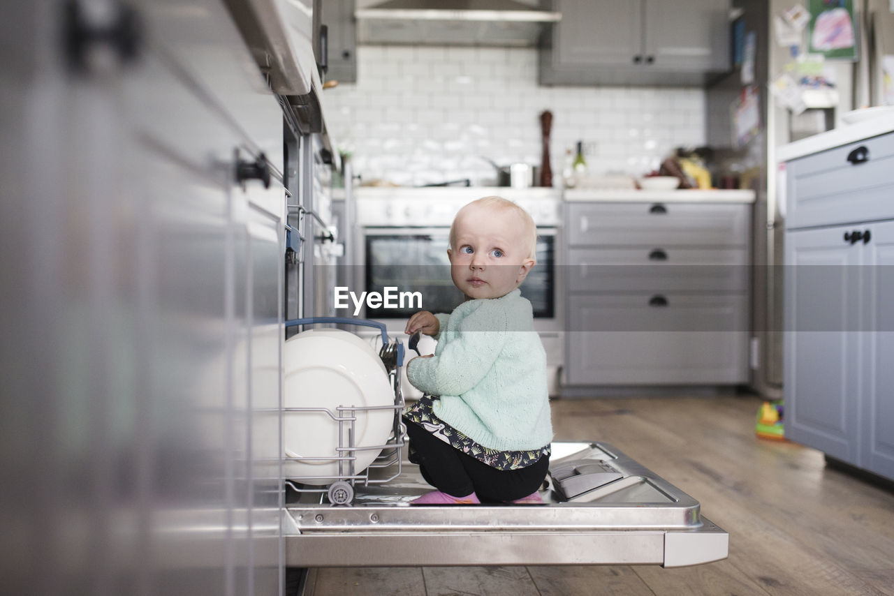 Cute girl looking away while sitting in dishwasher at kitchen