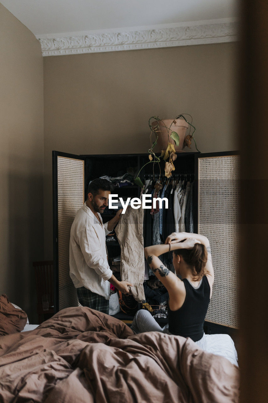 Man showing dress to non-binary person tying hair while sitting on bed at home