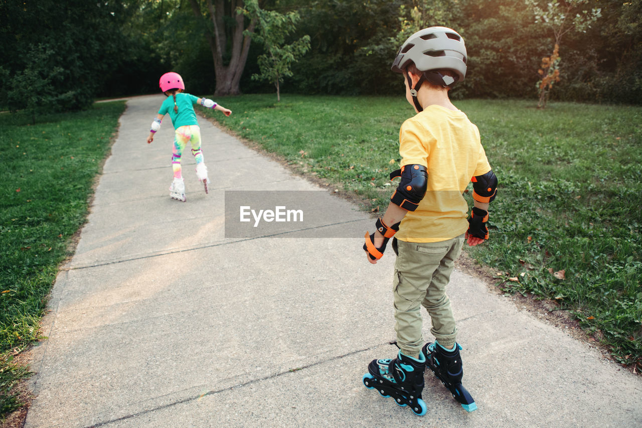 Friends boy and girl in helmets riding on roller skates in park on summer day. 