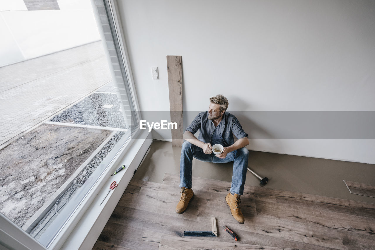 Mature man fitting flooring in new home, drinking coffee and taking a break