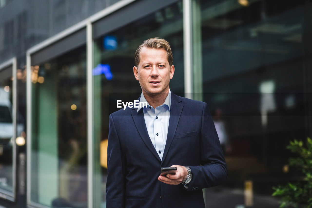 Portrait of mature businessman with smart phone standing against office building