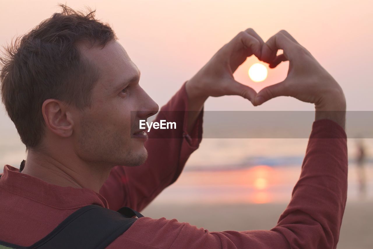 Side view of mid adult man making heart shape against sky during sunset