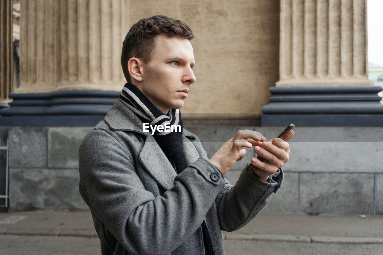 Portrait caucasian young man using his mobile phone in gray coat stands