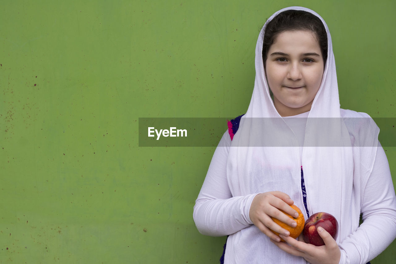 Portrait of smiling girl holding fruits while standing against green wall