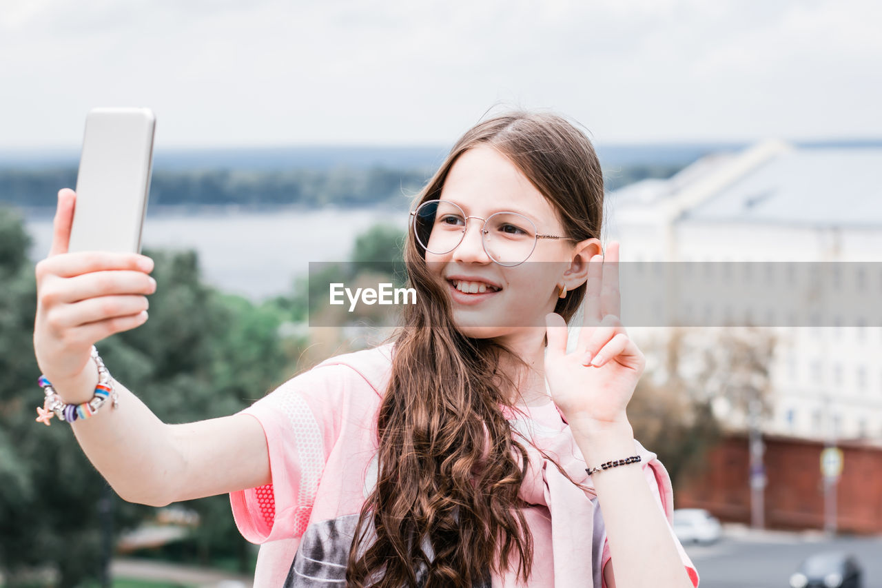 A cheerful girl in glasses takes a selfie on a smartphone on the background of a river and trees 