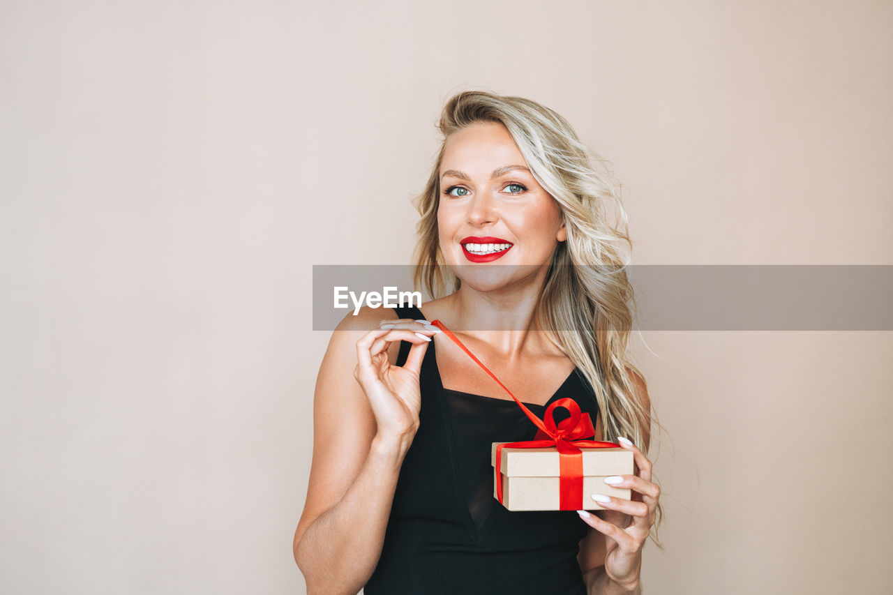 Portrait of woman with bright makeup in evening dress with gift box in hands on beige background