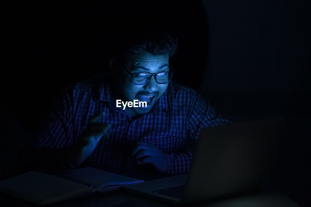 Young man screaming or shrieking at late night looking at laptop screen wearing eyeglass
