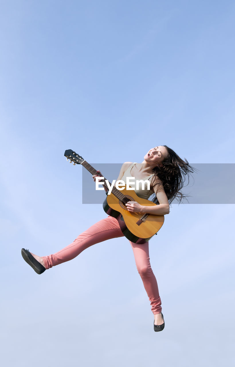 Low angle view of young woman playing guitar while jumping against clear blue sky
