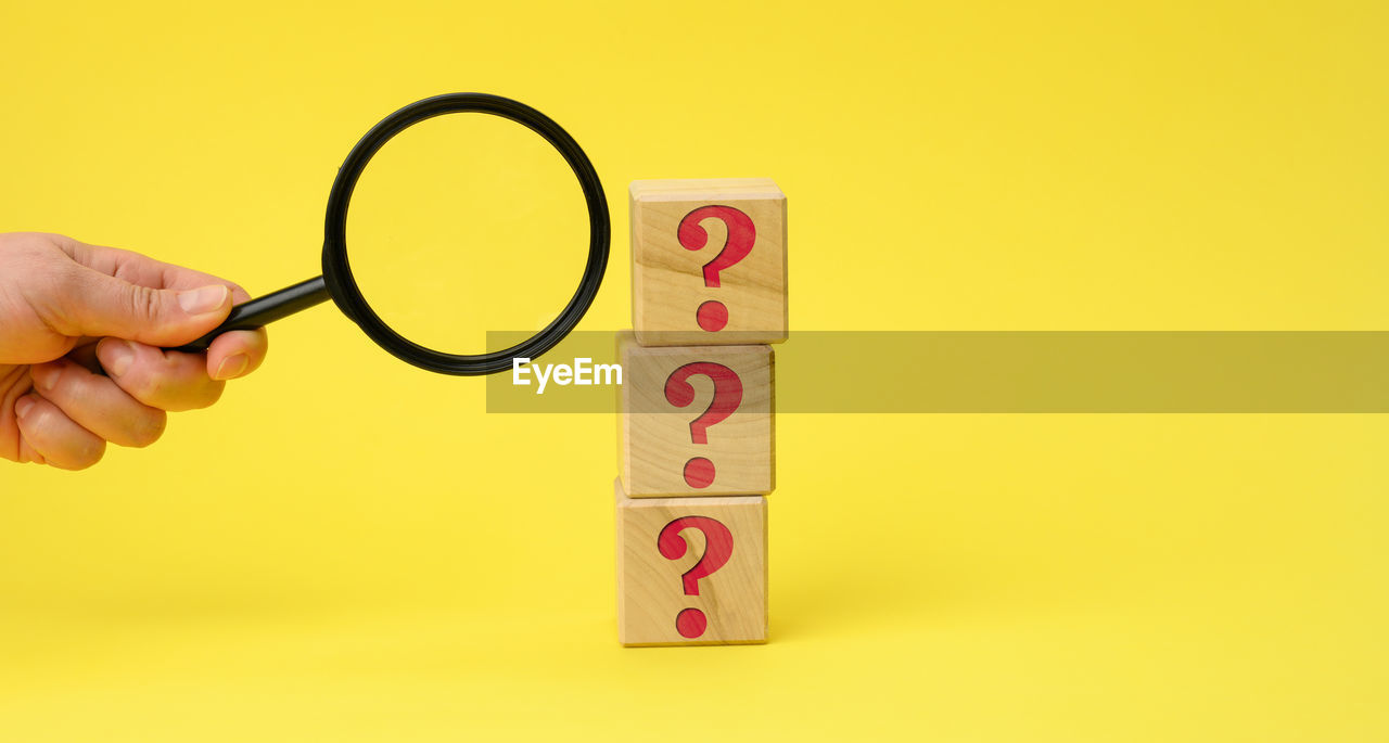 Wooden cubes with question marks and a female hand holds a magnifying glass on a yellow backgro